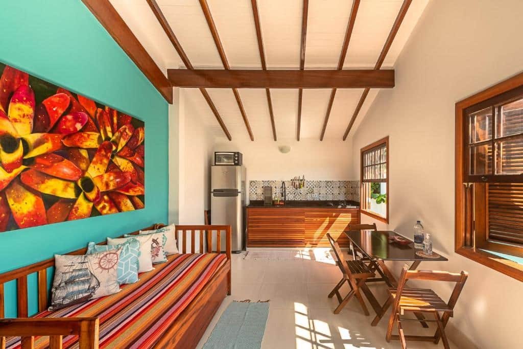 Do lado esquerdo um sofá cama e um quadro. No fundo uma pia e uma geladeira com um forno. Do lado direito uma mesa e duas janelas. Foto do Flats Vivendas do Mar para ilustrar post de airbnb em Paraty.