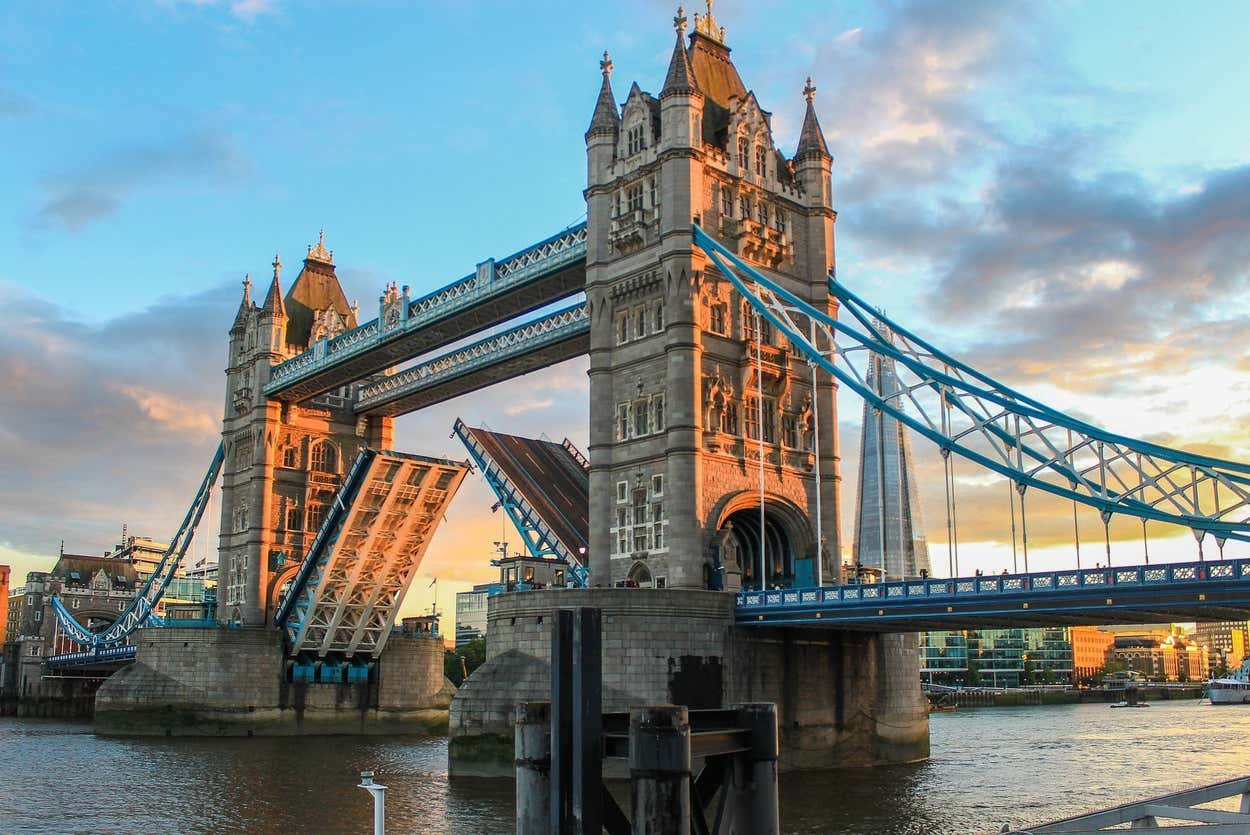 Tower Bridge em Londres com suas basculantes erguidas, iluminadas pelo pôr do sol, contra um céu parcialmente nublado e a paisagem urbana ao fundo.