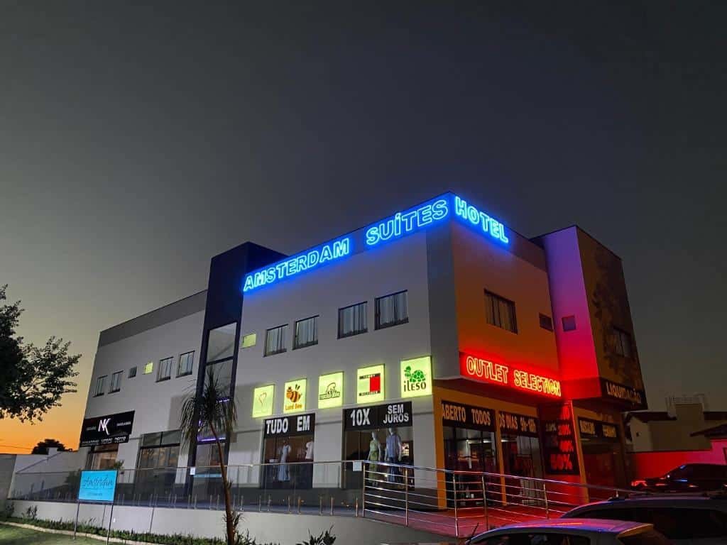 Vista a noite do Hotel Amsterdam Suítes, prédio de cor cinza com várias janelas, letreiros em neon azul com o nome do hotel em cima e abaixo lojas de roupas. Representa hotéis em Holambra.