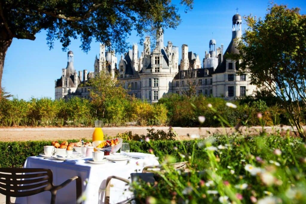 Frente do Relais de Chambord – Small Luxury Hotels of the World durante o dia com mesa de café a frente no meio do jardim ao fundo o castelo.