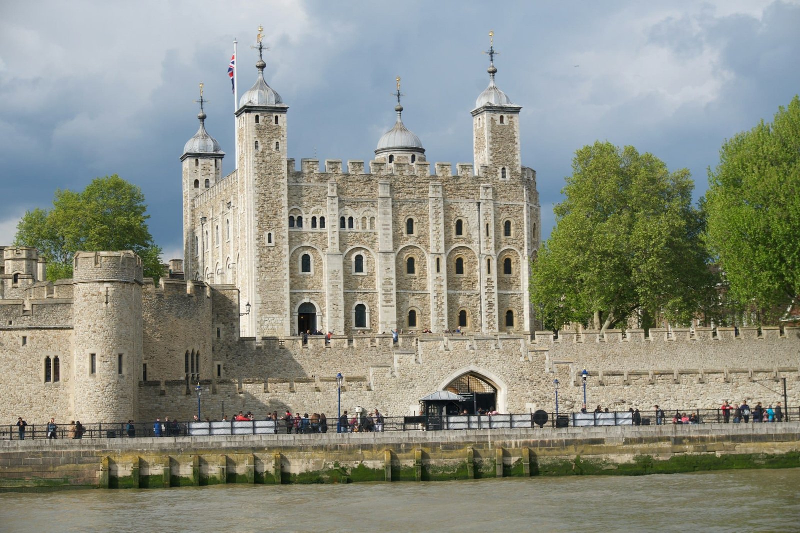 A Torre de Londres, um dos pontos turísticos de Londres, com seus muros de pedra, torres e bandeira, vista do outro lado do rio em um dia nublado.