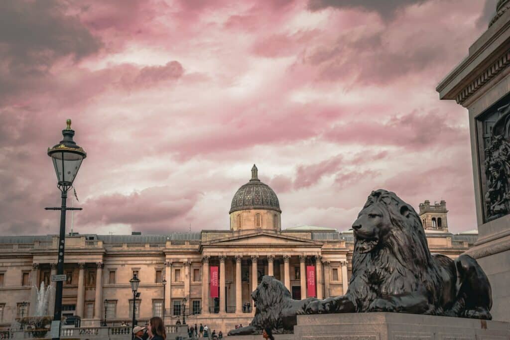 Vista da National Gallery com telhado abobadado e estátuas de leões na Trafalgar Square em primeiro plano, sob um céu nublado com tons de rosa.