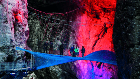 Pessoas em pé sobre uma ponte de rede azul dentro de uma caverna colorida em Glacier Canyon, com iluminação vermelha e azul iluminando paredes rochosas.
