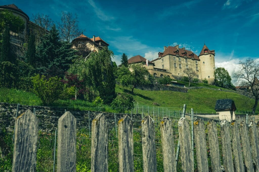 Imagem da cidade de Gruyères durante o dia com uma cerca a frente e ao fundo uma montanha e em cima dela do lado direito um castelo.