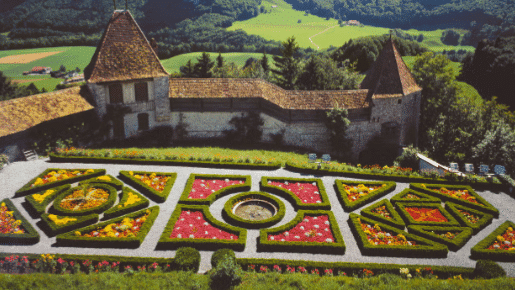 Vista aérea de um jardim formal  em Gruyères com padrões geométricos e diversas flores coloridas, cercado por árvores e muros de pedra.