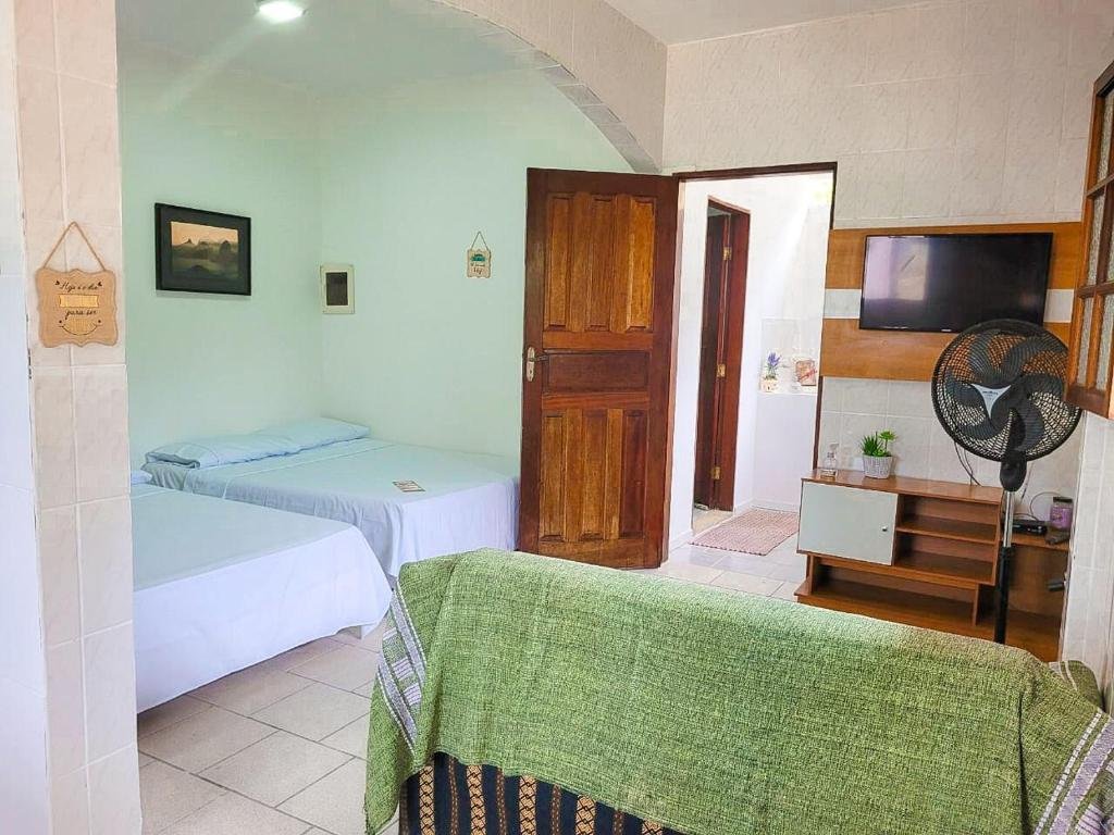 Quarto do Herdeiros do Pai, do lado esquerdo duas camas de casal, do lado direito um sofá, de frente um ventilador e uma raque com televisão, no fundo, no meio, uma porta.