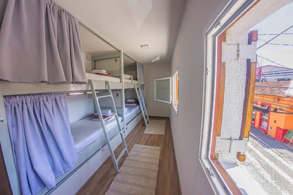 Um dos quartos do Hostel Central São Sebastião que mostra do lado esquerdo quatro beliches com cortinas de frente para duas janelas de vidro com vista para a rua. Imagem ilustrando post Pousadas pet friendly no Litoral Norte de São Paulo.