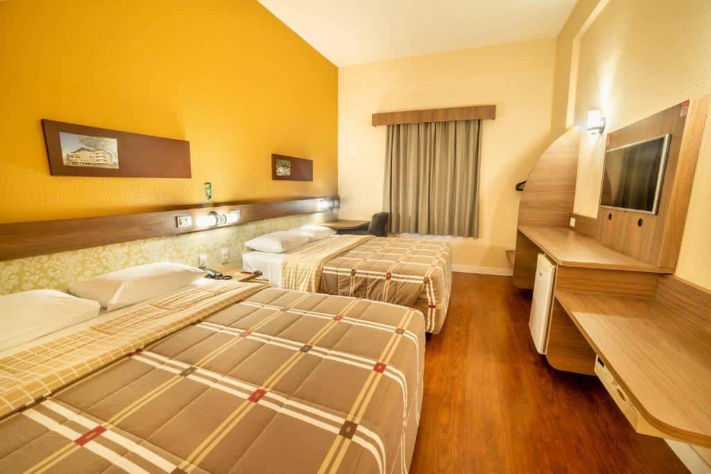quarto do Hotel 10 Joinville com duas camas de casal à esquerda e um grande painel de madeira em frente, com frigobar e uma televisão pendurada ao centro.