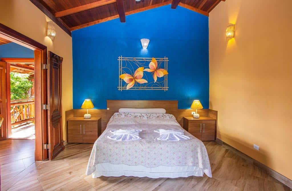 Hotel Alemão Beach de Ilhabela. Ilustra o post sobre hotéis em Ilhabela. O quarto tem paredes azuis e amarelas, com um teto de madeira escura.
Uma cama de casal com lençóis brancos está no centro do quarto. Duas toalhas dobradas em forma de cisne repousam sobre a cama.
Mesinhas de cabeceira de madeira com luminárias acesas estão posicionadas de cada lado da cama.
Na parede azul atrás da cama, há uma arte decorativa feita de metal, representando folhas e um quadro geométrico.
O piso é composto por tábuas largas de madeira escura.
À esquerda da imagem, portas duplas abertas revelam um espaço exterior iluminado pelo sol.