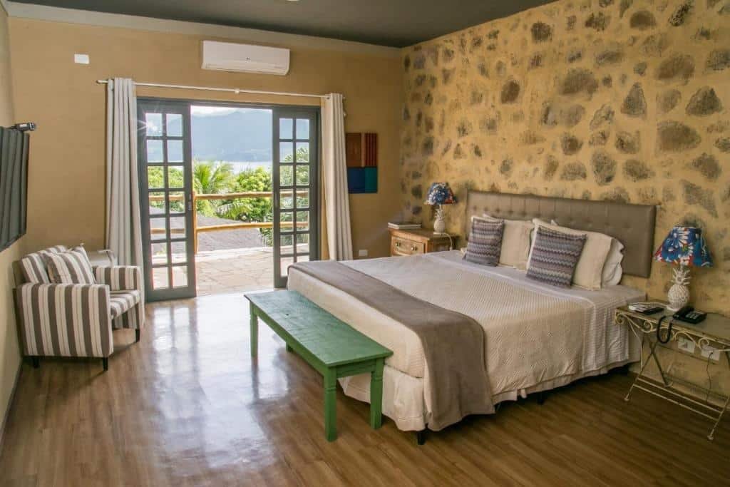 Hotel Boutique Ananas. Ilustra o post sobre hotéis em Ilhabela. A foto mostra um quarto bem iluminado com uma cama, móveis e uma varanda que oferece uma vista para as montanhas. O quarto tem paredes de pedra e piso de madeira, dando-lhe um toque rústico e acolhedor. Há uma cama grande no centro do quarto com uma colcha clara e almofadas decorativas. Uma mesa de cabeceira branca com detalhes ornamentados está ao lado da cama, segurando um abajur azul decorativo. À esquerda da imagem, há uma poltrona listrada confortável com um design moderno. Uma grande janela de vidro fornece muita luz natural ao quarto e abre para uma varanda com vista para as montanhas verdes ao fundo. Um banco verde longo está localizado na frente da cama.