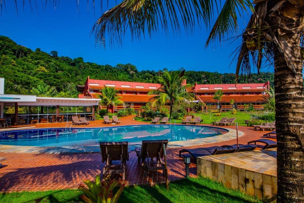 Parte do Hotel Boutique Frangipani que mostra uma grande piscina azul com algumas cadeiras e espreguiçadeiras em volta. Ao fundo tem a construção do hotel e em volta há bastante árvores verdes durante o dia, ilustrando post Airbnb em Brotas.