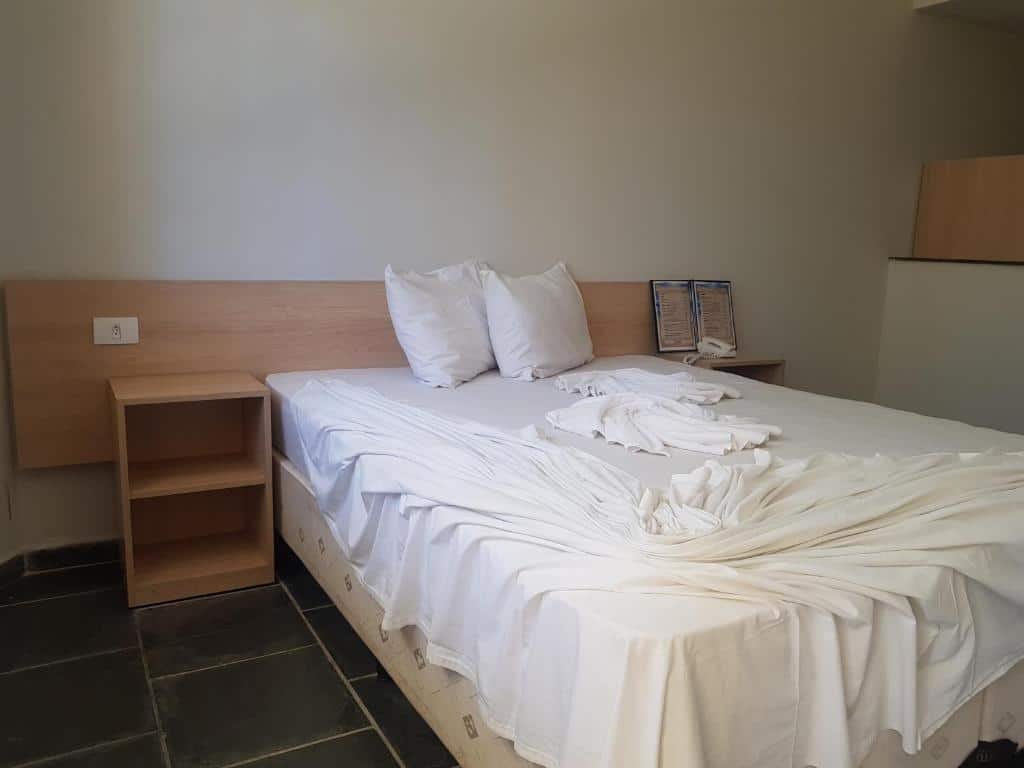 Hotel da Ilha. Ilustra o post sobre hotéis em Ilhabela. A imagem mostra uma cama de casal com lençóis brancos desarrumados.
Dois travesseiros estão encostados na cabeceira da cama.
A cabeceira da cama é de madeira clara e combina com um pequeno criado-mudo posicionado à esquerda da cama.
No criado-mudo, há um telefone fixo e alguns papéis ou panfletos organizados em um suporte.
O piso é feito de lajes escuras, criando contraste com as paredes e a roupa de cama clara.
Uma tomada elétrica está visível na parede acima do criado-mudo.