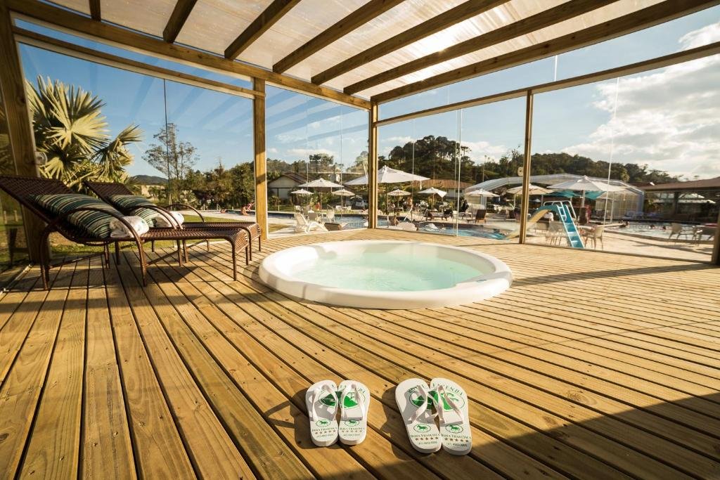 Deque de madeira coberto com hidromassagem no Hotel Fazenda Dona Francisca, em Joinville. Ao fundo é possível ver uma piscina e várias cadeiras com guarda-sol sob um céu azul claro