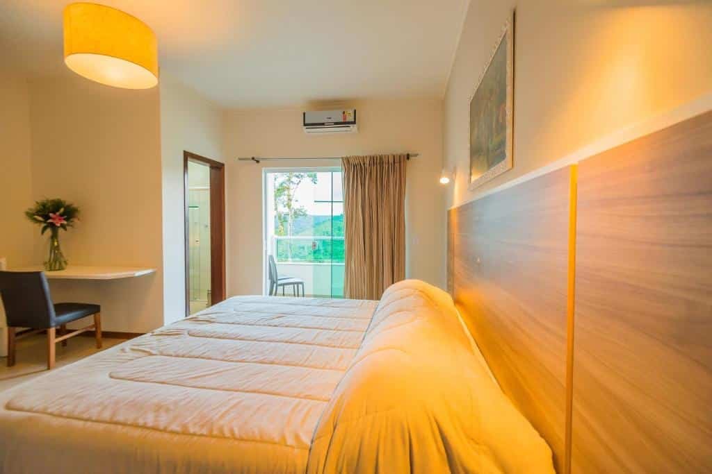 Quarto do Hotel Fazenda Dona Francisca com uma cama de casal à direita e uma sacada com a porta aberta ao fundo do quarto. A vista é para uma área verde com montanhas