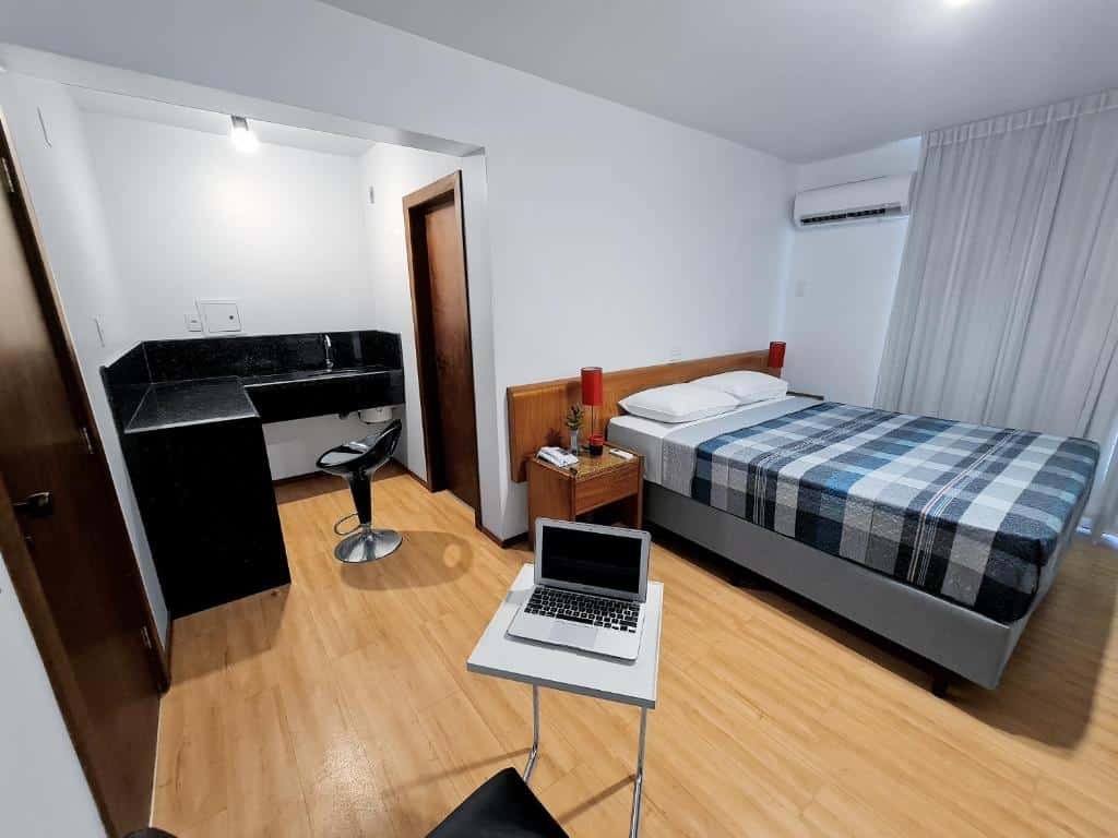 Quarto do Germânia, com uma cama de casal à direita do quarto, com uma mesa pequena e um computador em cima, ao lado da cama, na parte inferior da imagem. Há também um balcão ao lado da porta do banheiro, no canto esquerdo do quarto.