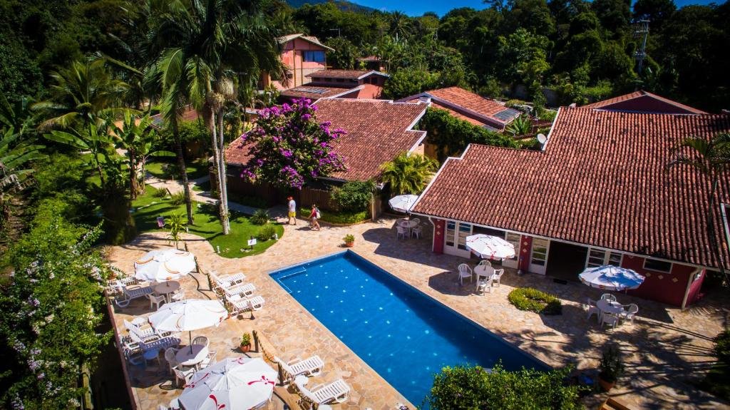 Hotel Ilhasol. A imagem apresenta uma vista aérea de um ambiente externo ensolarado e verdejante que parece ser um resort ou uma pousada.
Há uma piscina retangular azul clara no centro da imagem, cercada por espreguiçadeiras brancas e mesas com guarda-sóis.
Edifícios: Existem edifícios com telhados vermelhos; o design arquitetônico sugere um estilo tropical ou costeiro. A vegetação é exuberante, com várias palmeiras e outras árvores tropicais espalhadas pelo local. Há também flores roxas visíveis crescendo em arbustos ou árvores próximas aos edifícios. O céu é claro e azul, indicando um dia ensolarado e agradável.