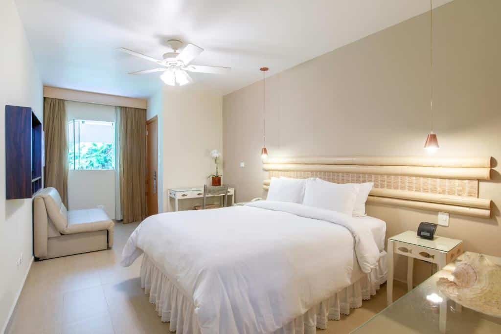 Hotel Itapemar. Ilustra o post sobre hotéis em Ilhabela. O quarto é espaçoso e possui paredes claras, o que contribui para uma atmosfera luminosa.
Há uma cama grande no centro do quarto com lençóis brancos e travesseiros.
Acima da cama, há uma cabeceira longa com iluminação embutida que proporciona uma luz suave ao ambiente.
Do lado direito da cama, há um criado-mudo com um telefone em cima.
À esquerda da imagem, há uma poltrona branca encostada na parede abaixo de um quadro azul escuro.
Uma janela grande está localizada à esquerda do quarto, coberta por cortinas bege pesadas. Através dela pode-se ver a vegetação externa.
Um ventilador de teto branco está instalado no teto, ajudando na circulação do ar dentro do quarto.