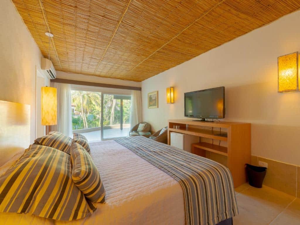 Hotel Mercedes. Ilustra o post sobre hotéis em Ilhabela. A foto mostra um quarto de hotel bem iluminado e aconchegante. Aqui estão os detalhes:

O quarto é espaçoso e possui duas camas de solteiro com lençóis listrados em tons de amarelo, cinza e branco.
As camas estão arrumadas, e cada uma tem um travesseiro.
Há uma grande janela ao fundo que permite a entrada de muita luz natural, revelando uma visão das palmeiras do lado de fora.
O teto é coberto com um material trançado marrom claro, dando um toque rústico ao ambiente.
Na parede à direita, há uma TV plana montada acima de um móvel de madeira que parece ter alguns compartimentos para armazenamento.
Duas luminárias quadradas estão fixadas na parede em ambos os lados da janela, emitindo uma luz suave.
Um aparelho de ar condicionado está instalado no canto superior esquerdo da imagem.