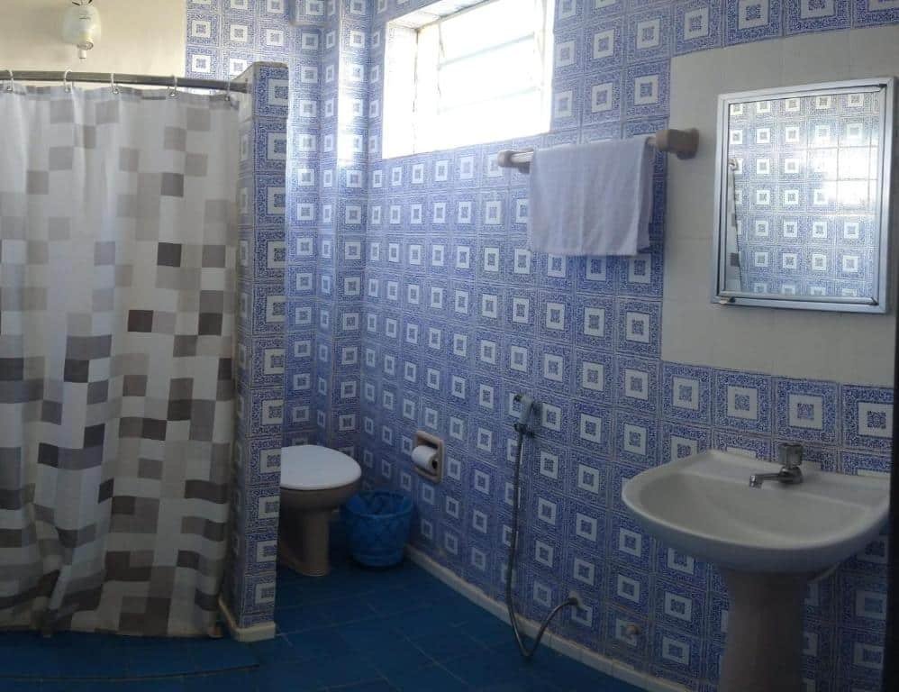 Foto da Hotel Pousada Icaraí.  Nessa foto, há um banheiro com azulejos azuis e brancos nas paredes, um chão azul, uma cortina de chuveiro cinza e branca, um vaso sanitário, uma pia branca com torneira prateada, uma lixeira azul e toalhas. A janela está na parede oposta à entrada.