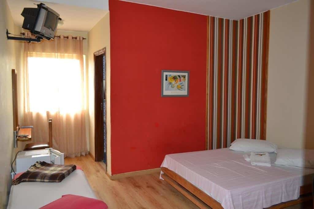 Foto da Hotel Pousada Icaraí que está ilustrando o post de pousadas pet friendly em Minas Gerais. Nessa foto, há um quarto com uma cama de casal à direita, coberta por uma colcha branca e um travesseiro. À esquerda da cama, há uma pequena mesa de cabeceira com um telefone. Uma segunda cama ou sofá-cama está parcialmente visível no canto inferior esquerdo. A parede atrás da cama é vermelha com uma pintura pendurada. Há também uma parede listrada à direita. Um projetor está montado no teto. A luz do dia entra pela janela à esquerda, que tem cortinas puxadas para os lados.