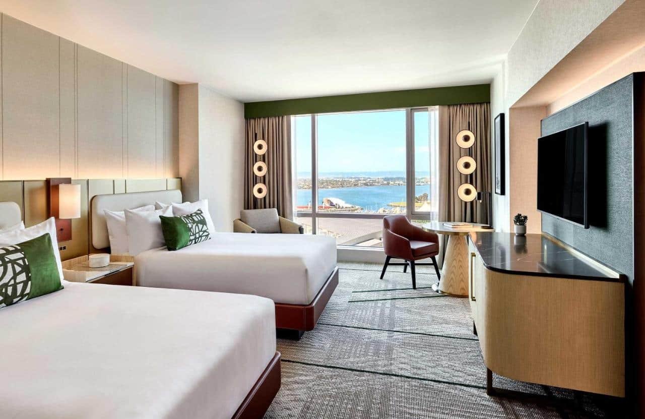 Quarto do Omni San Diego Hotel. Do lado esquerdo duas camas de solteiro, entre elas uma cômoda e no fundo uma poltrona. Do lado direito uma cômoda com TV, no fundo uma mesa de trabalho. No fundo, uma parede de vidro com vista para o mar.