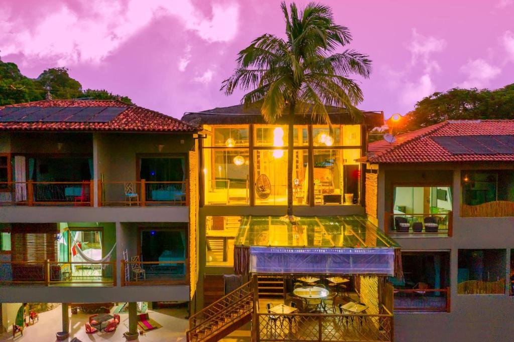 Foto do Hotel Vila Kebaya mostrando seu exterior. O céu no fundo está na cor violeta. O prédio no centro está iluminado com luzes laranjas. Vemos as varandas de capa apartamento.