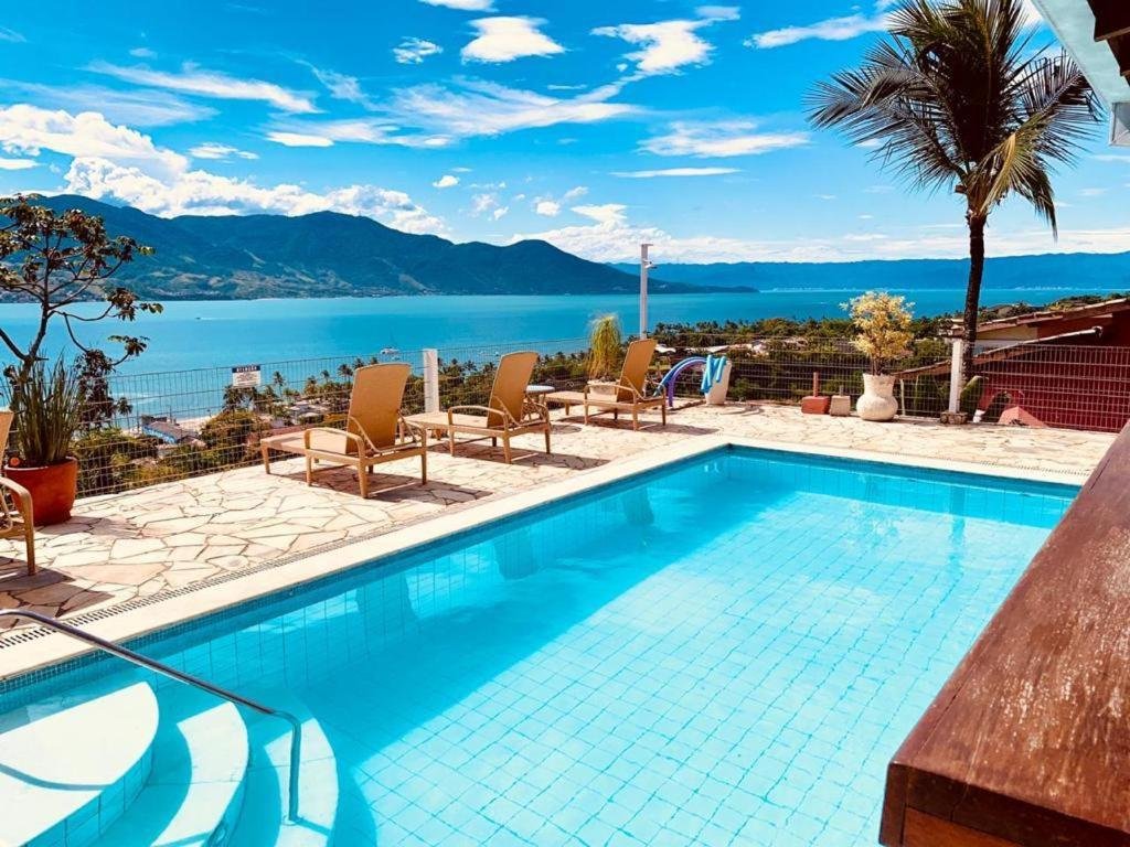 Hotel Vista Bella. Há uma piscina com uma vista deslumbrante para um lago e montanhas. Vamos explorar os detalhes:

A imagem mostra uma piscina de água azul cristalina, cercada por um deck de pedra.
Há quatro espreguiçadeiras alinhadas ao lado da piscina, voltadas para a vista panorâmica do lago e das montanhas ao fundo.
Uma palmeira e outras plantas estão próximas à área da piscina, adicionando um toque tropical à cena.
O céu está claro com algumas nuvens esparsas, indicando um dia ensolarado e agradável.
As montanhas ao fundo são majestosas e parecem cercar o lago, criando uma atmosfera serena.
Uma cerca baixa separa a área da piscina da vista espetacular, garantindo segurança sem obstruir a visão.