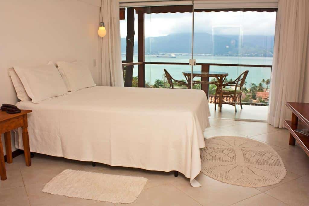 Hotel Vista Bella. Ilustra o post sobre hotéis em Ilhabela. O quarto é espaçoso e bem iluminado, com piso de azulejos claros.
Há uma cama grande com lençóis brancos e travesseiros no canto esquerdo do quarto.
Ao lado da cama, há um criado-mudo de madeira escura com um telefone preto em cima.
No chão, perto da cama, há um tapete retangular branco pequeno e outro tapete redondo maior com um design geométrico.
Há uma grande janela panorâmica que oferece uma vista deslumbrante do oceano, montanhas ao fundo e céu nublado.
Na frente da janela, há uma mesa de madeira escura com duas cadeiras que oferecem um local para sentar e apreciar a vista.