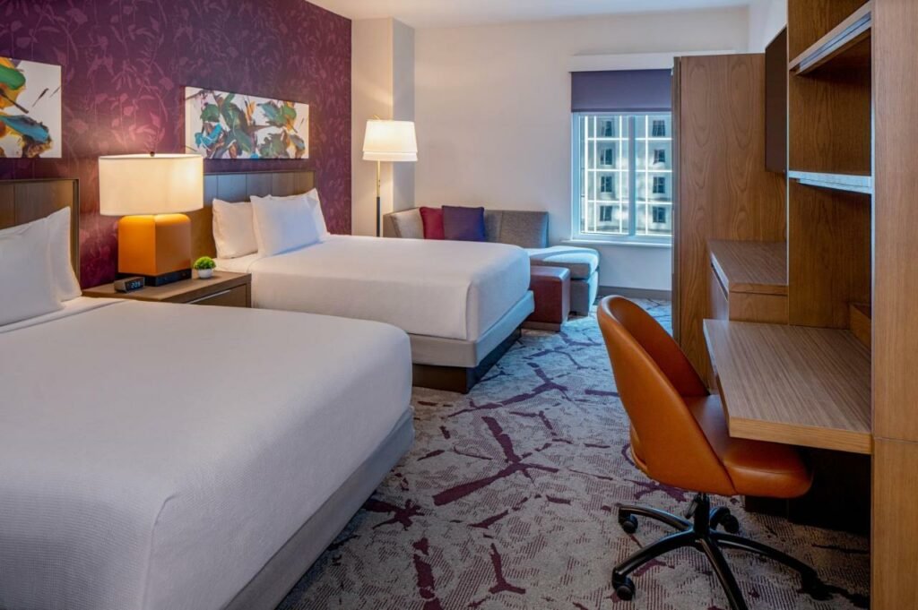 Quarto do Hyatt House Tampa Downtown com uma cama de solteiro, uma de casal, um sofá, uma janela, um carpete e uma bancada de madeira com uma cadeira giratória