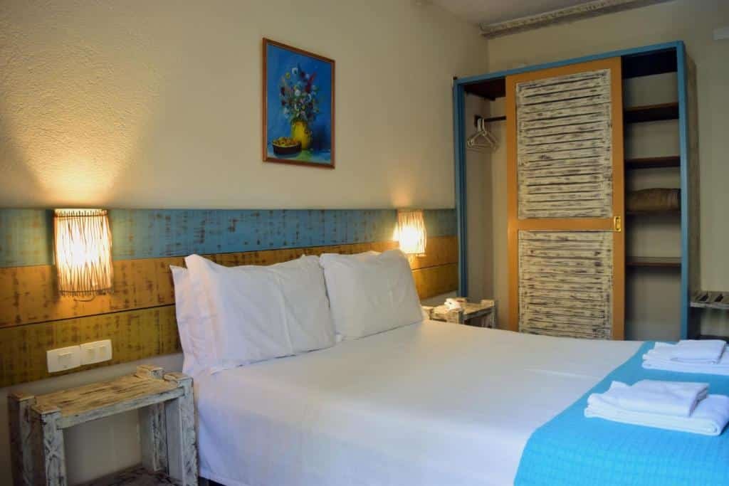 Ilha Deck Hotel. Ilustra o post sobre hotéis em Ilhabela. O quarto é iluminado e apresenta paredes claras.
Há uma cama grande no centro com lençóis brancos e uma colcha azul parcialmente estendida sobre ela.
Duas mesinhas de cabeceira rústicas flanqueiam a cama, cada uma com uma luminária pendurada acima.
Acima da cama, há um quadro colorido na parede que parece representar flores em um vaso.
À direita da imagem, há um armário embutido com portas de persiana para ventilação.