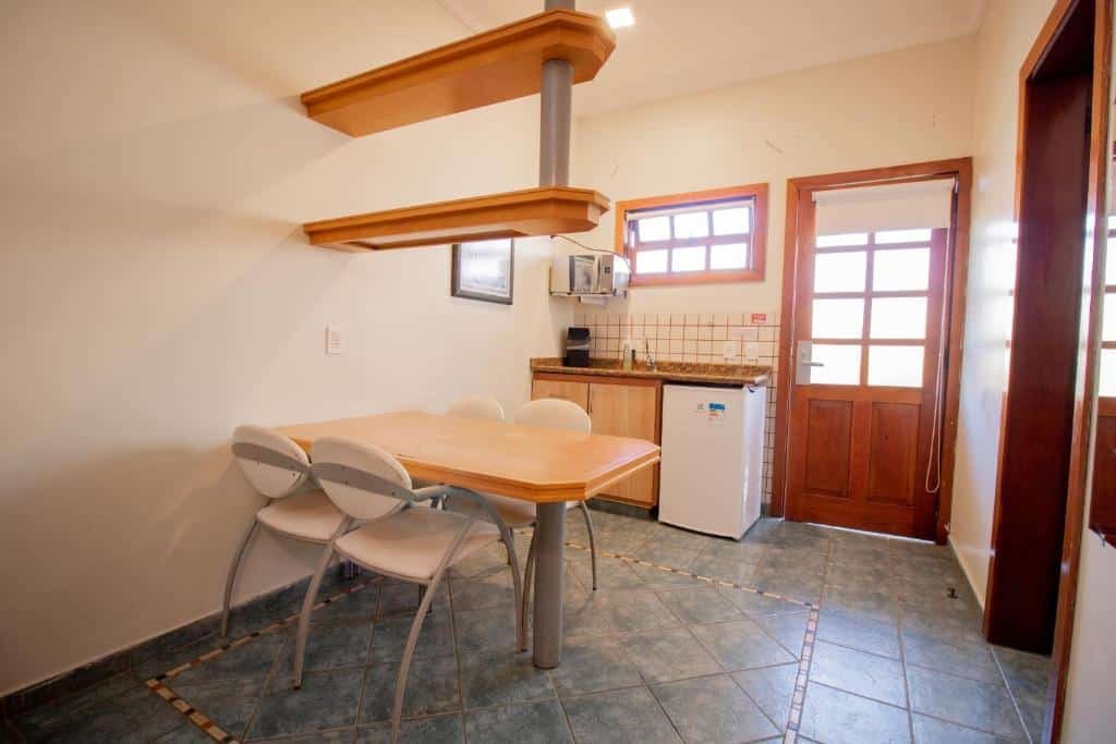Cozinha do Ilha Flat Hotel. Uma mesa com quatro cadeiras está em frente à bancada da pia com frigobar e micro-ondas. Em cima da pia está uma janela, e logo ao lado fica a porta.