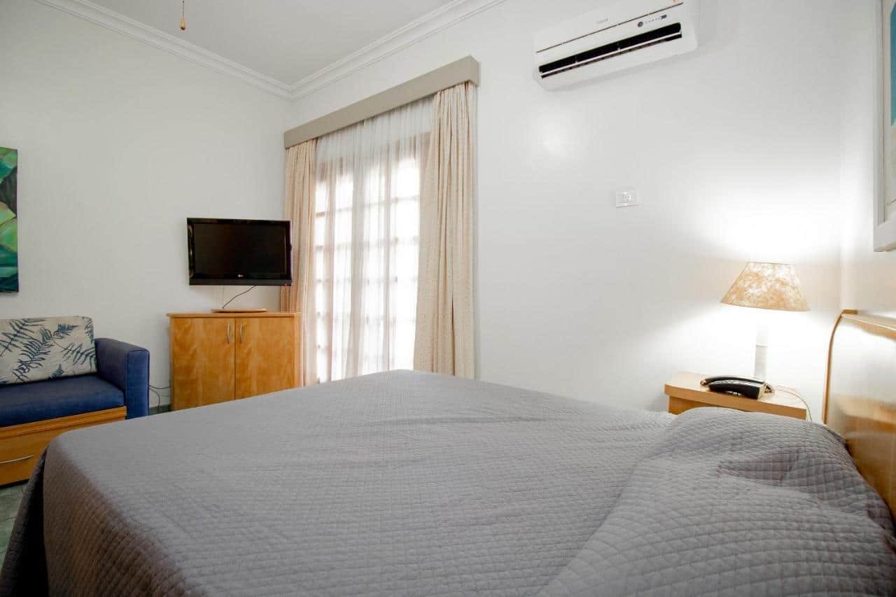 Quarto do Ilha Flat Hotel, um dos airbnb em Ilhabela. Uma cama de casal encostada na parede ao lado direito tem mesinhas de cabeceira com abajur dos dois lados e encara um sofá e uma cômoda com TV na parede oposta. Na lateral direita da cama há um aparelho de ar-condicionado ao lado de uma janela com cortinas.