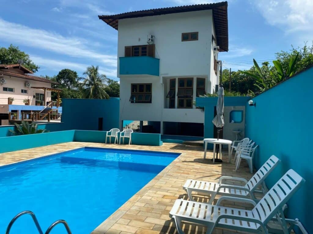 Piscina da Ilhabela Casa com Piscina rodeada por espreguiçadeiras e mesinhas com cadeiras e guarda-sóis. Ao fundo está o sobrado.