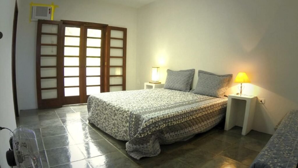 Quarto da Ilhabela Casa com Piscina, um dos airbnb em Ilhabela. Uma cama de casal com mesinhas de cabeceira e abajures dos dois lados está encostada na parede central. Na lateral esquerda da cama fica a porta e o aparelho de ar-condicionado, já na lateral direita está uma cama de solteiro. Na parede em frente à cama há um ventilador.