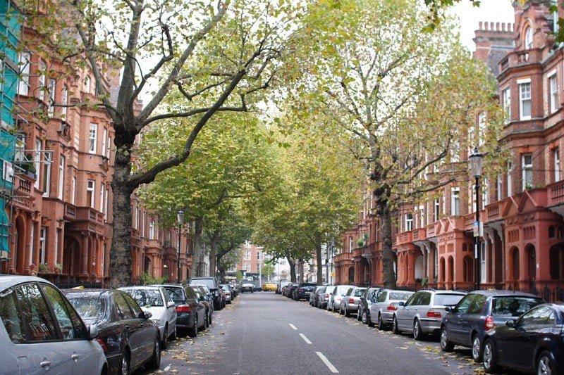 Rua arborizada no bairro de Chelsea com carros estacionados e prédios de tijolos vermelhos em ambos os lados sob um céu parcialmente nublado. A imagem ilustra o post sobre roteiro para Londres.