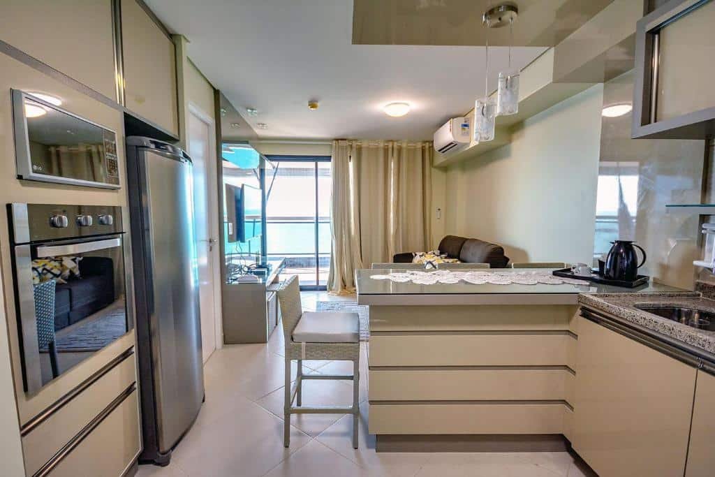 Cozinha do Landscape Beira Mar Platinum. Do lado esquerdo está o fogão, o micro-ondas e uma geladeira. Do lado direito está a pia e o balcão com uma cadeira. Ao fundo está a sala de estar com um sofá e uma tv. Ao lado da sala está com uma varanda. 