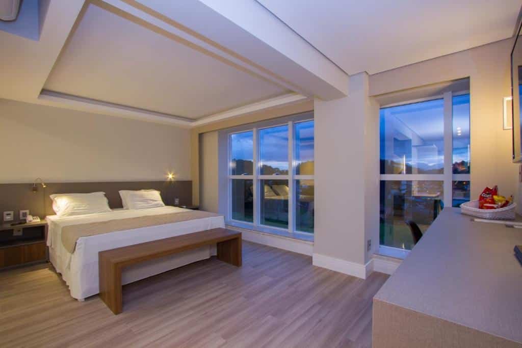 Quarto do Le Canard, com 1 cama de casal grande à esquerda e grandes janelas de vidro à direita. No canto direito da imagem há um balcão