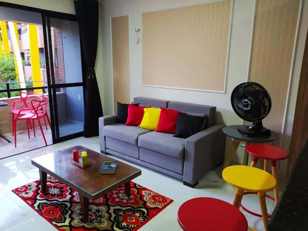 Sala de estar do Lindo Apartamento na Praia do Futuro. Ao fundo está a varanda com cadeiras e uma mesinha. Na sala um sofá está encostado na parede de frente para uma mesinha de centro e um tapete. Ao lado do sofá está um ventilador e alguns banquinhos.