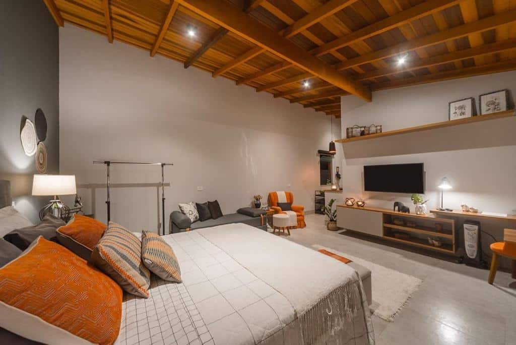 Quarto do Loft Luz. Uma cama de casal do lado esquerdo com almofadas, no fundo uma arara e um sofá cama com uma poltrona. Do lado direito uma raque e uma televisão. Foto para ilustrar post sobre airbnb em Santo Antônio do Pinhal.