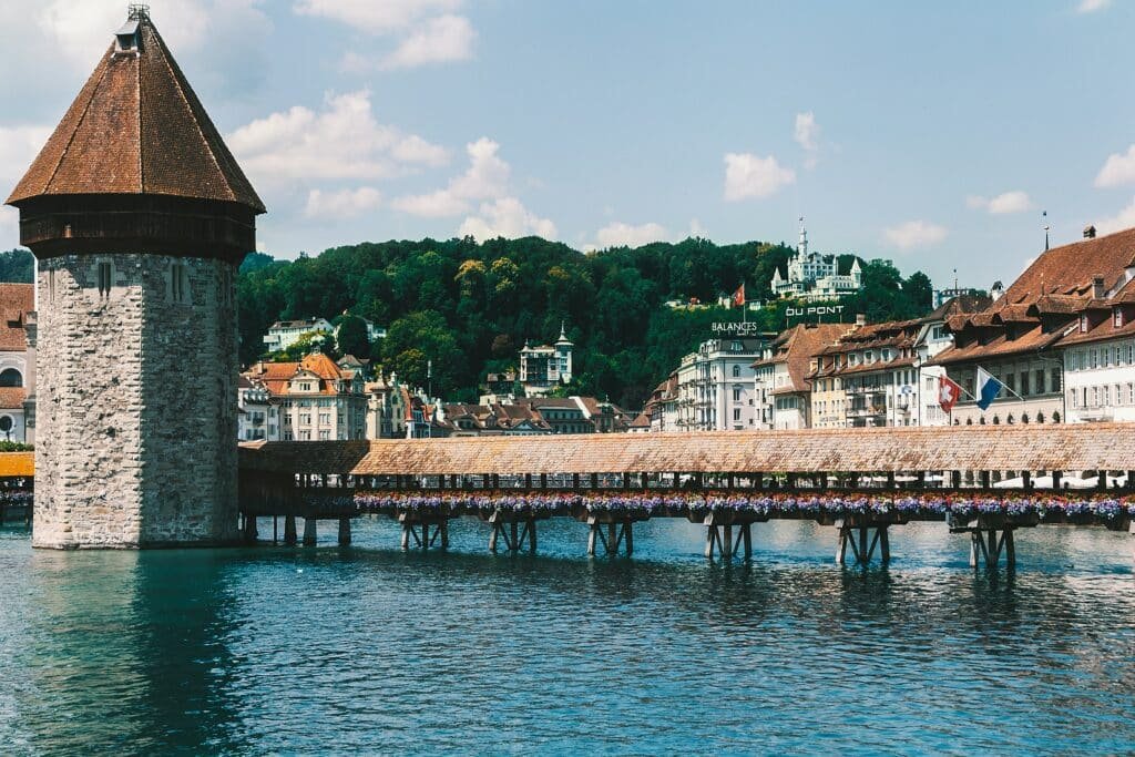 Imagem da cidade de Lucerna durante o dia com rio do lado direito a frente e no centro uma ponte que que vai até uma torre que está do lado esquerdo e ao fundo vários prédios. Representa o que fazer na Suíça.