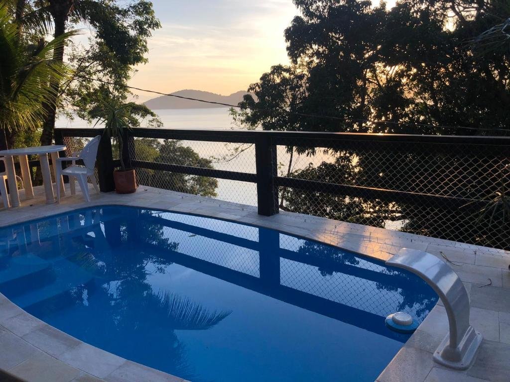 Piscina da Mansão na costa com vista para o mar. No fundo mesa e cadeira, um cercado com vista para as árvores e o mar.