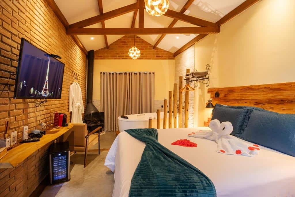 Quarto do Mantiqueira Flat. Uma cama de casal do lado direito, no fundo uma banheira de hidromassagem. Do lado esquerdo uma estante, uma televisão, uma poltrona e uma lareira. Foto para ilustrar post sobre airbnb em Santo Antônio do Pinhal.