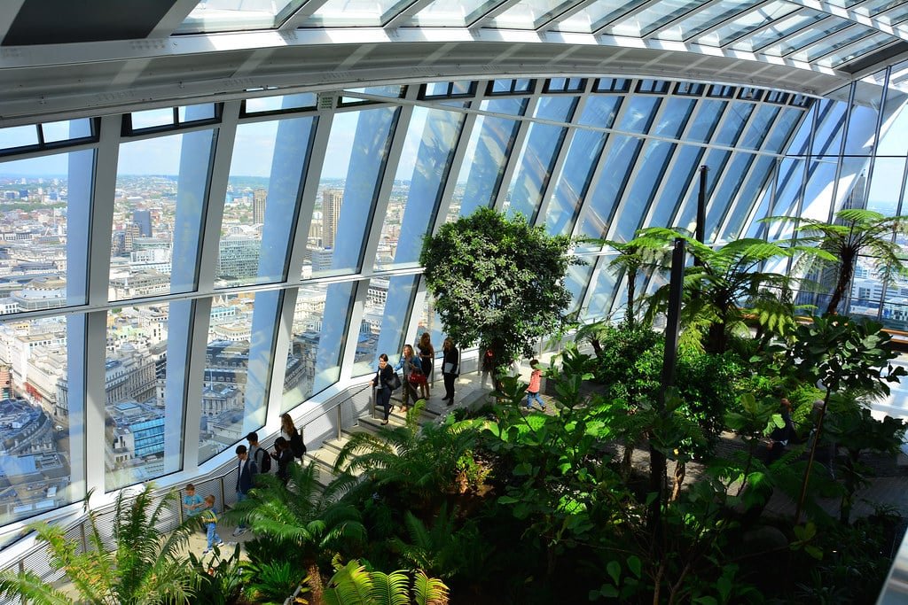Pessoas caminhando por um jardim interno com grandes janelas com vista para a paisagem urbana. Esse é o Sky Garden, uma das opções de o que fazer em Londres.