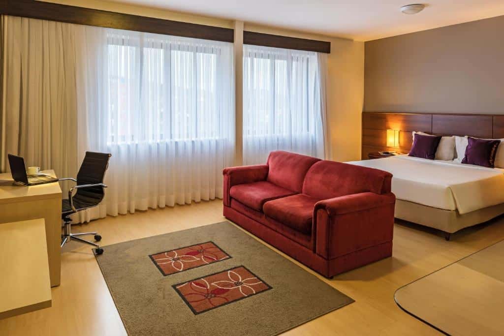 Quarto do Mercure Joinville Prinz com um sofá vermelho em frente a cama de casal e com uma mesa de trabalho na frente do sofá.