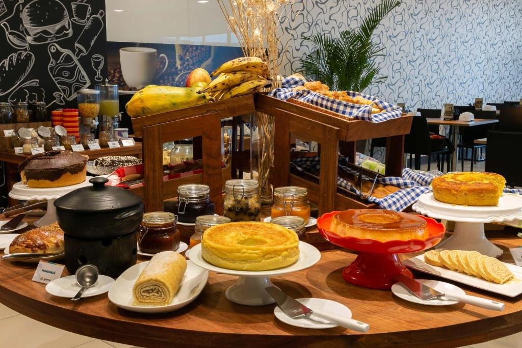 Mesa redonda no meio do saguão do WA Hotel Caruaru. Na mesa estão diversos alimentos preparados para o café da manhã como bolo de rolo, frutas, pudim e bolos.