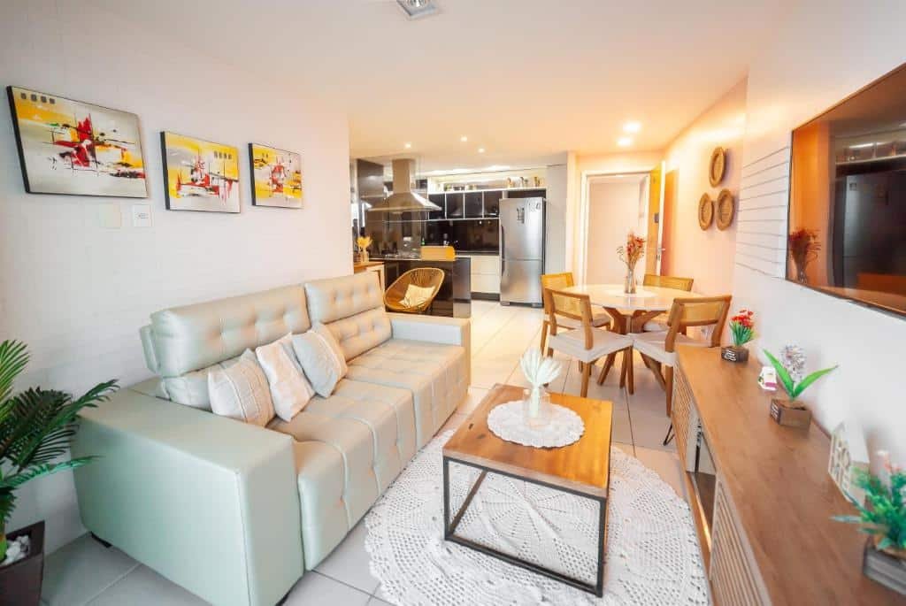 Sala de estar do Modern Beachfront Apartment w. Magnificent Seaview. Um sofá está de frente para a tv e entre eles está uma mesinha de centro e um tapete. Ao fundo há uma mesa redonda de quatro lugares. Mais ao fundo está a cozinha com geladeira, armários, balcão e cadeiras.