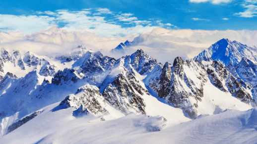Picos de montanhas cobertos de neve sob um céu azul claro com nuvens dispersas ao fundo.
