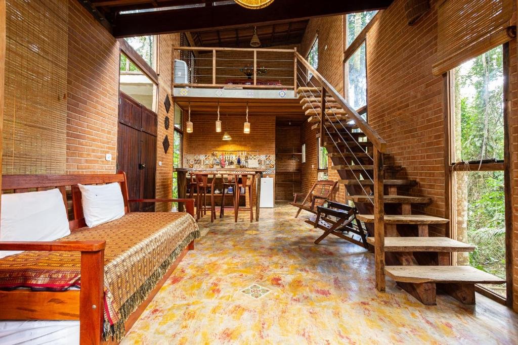 Oca Paraty Chalé Ecológico. Do lado direito uma escada de madeira e janelas de vidro, do lado esquerdo um sofá cama e a porta do chalé. No fundo um balcão de madeira com luminárias e cadeiras de madeira.