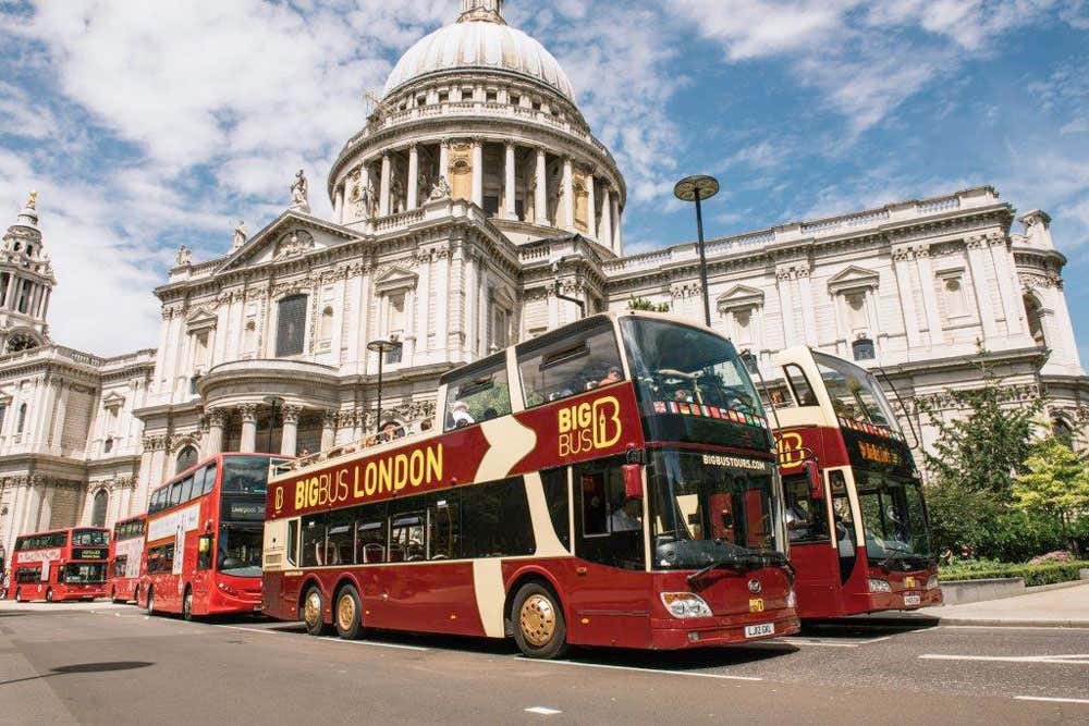 Dois ônibus de turismo vermelhos de dois andares em frente a um edifício branco ornamentado com uma grande cúpula sob um céu parcialmente nublado.