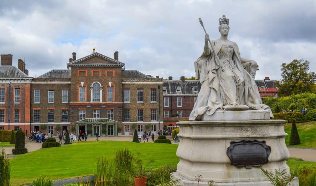 Estátua de uma rainha sentada com um cetro em frente ao Palácio de Kensington, cercada por jardins e visitantes sob um céu nublado.